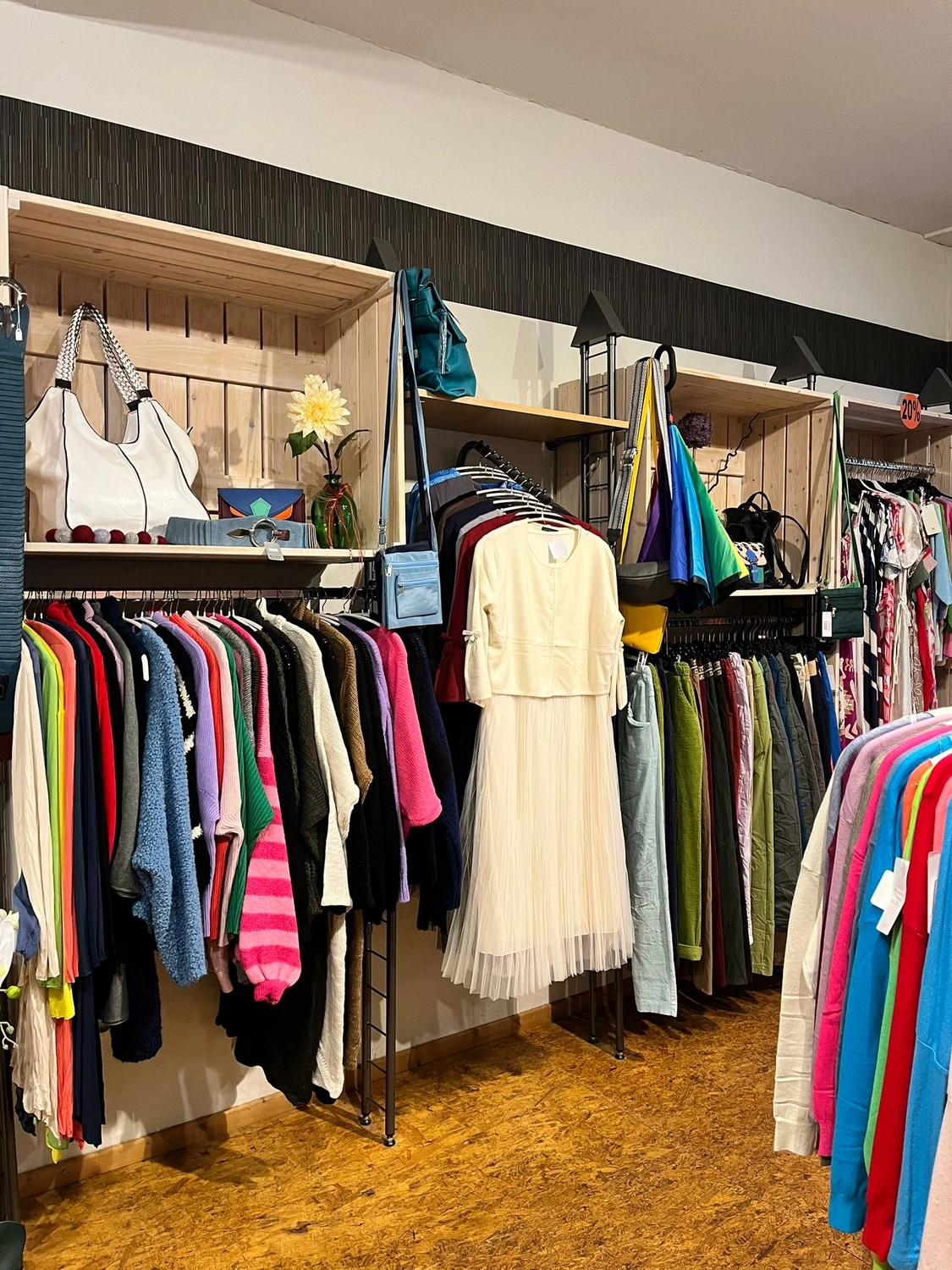 Mode & Handarbeitsdiele - Wolle, Kunsthandwerk, Mode und Accessoires - Ein Verkaufsdisplay mit einem weißen Kleid, das zwischen verschiedenen farbenfrohen Oberteilen und Handtaschen in Regalen hängt.