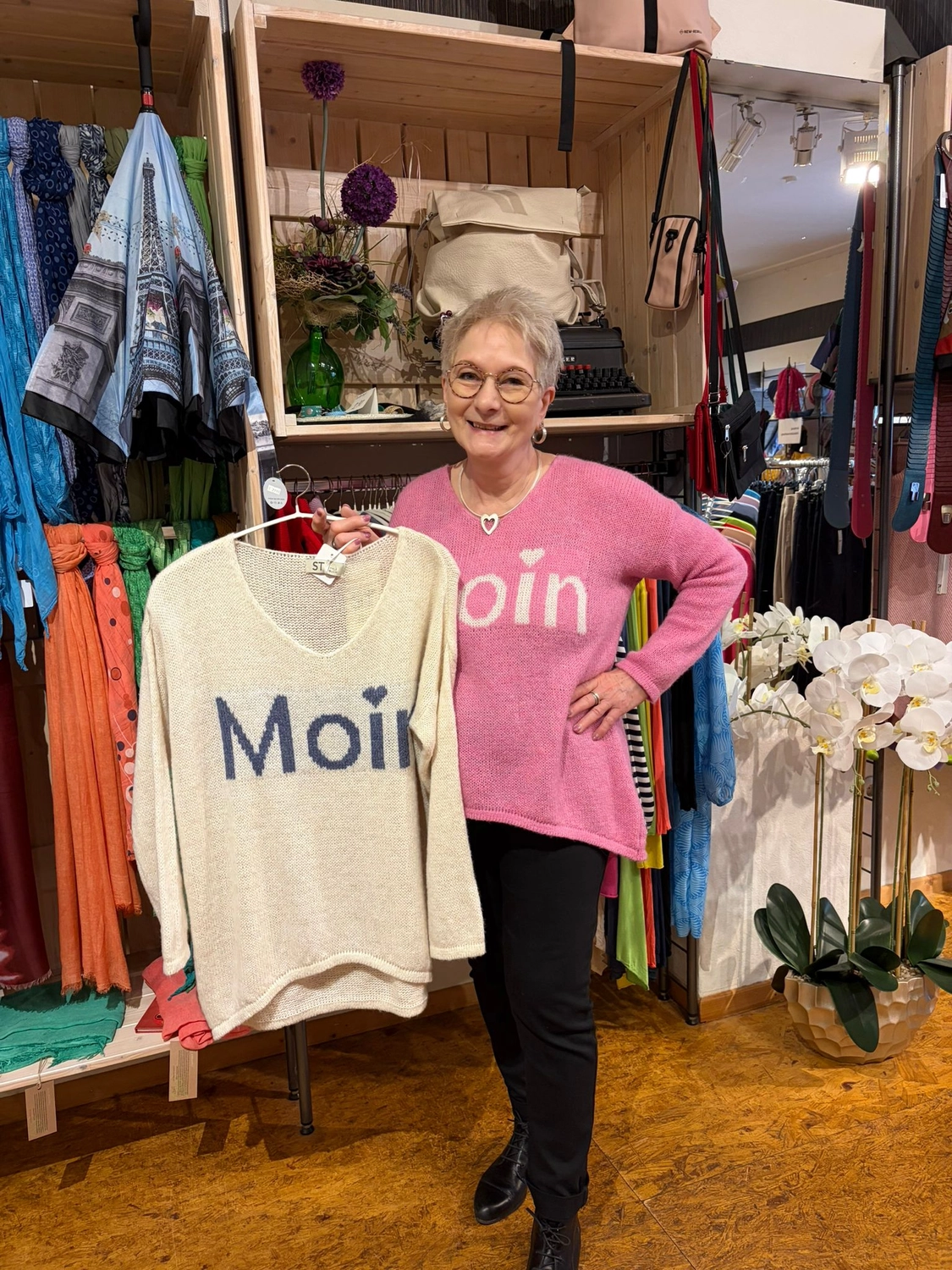 Mode & Handarbeitsdiele - Wolle, Kunsthandwerk, Mode und Accessoires - Eine Frau in einem pinkfarbenen „Moin“-Pullover hält in einer Modeboutique einen ähnlichen cremefarbenen Pullover hoch.