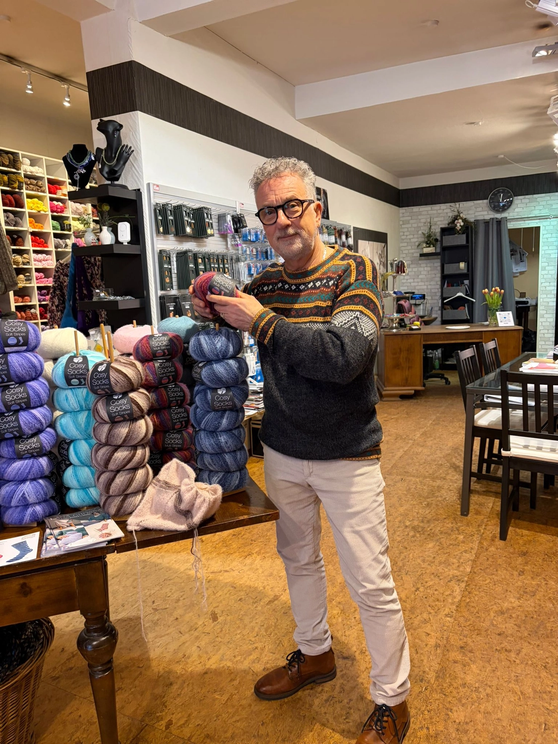 Mode & Handarbeitsdiele - Wolle, Kunsthandwerk, Mode und Accessoires - Ein Mann in einem gemusterten Pullover und mit Brille steht in einem Bastelladen neben Stapeln bunter Wolle.