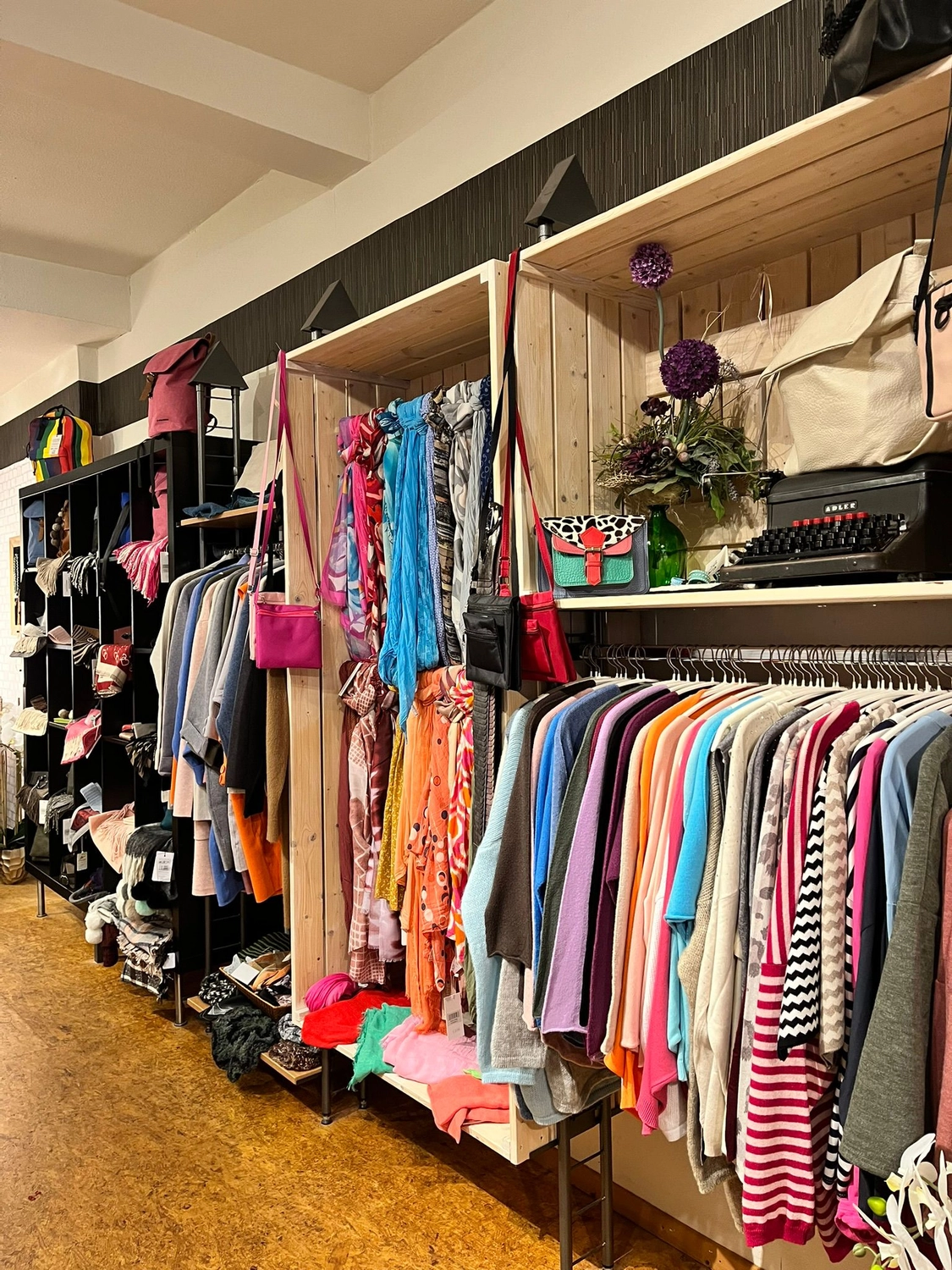 Mode & Handarbeitsdiele - Wolle, Kunsthandwerk, Mode und Accessoires - Blick in den Ladeninnenraum mit Ständern voller farbenfroher Kleidung und Accessoires an der Wand.