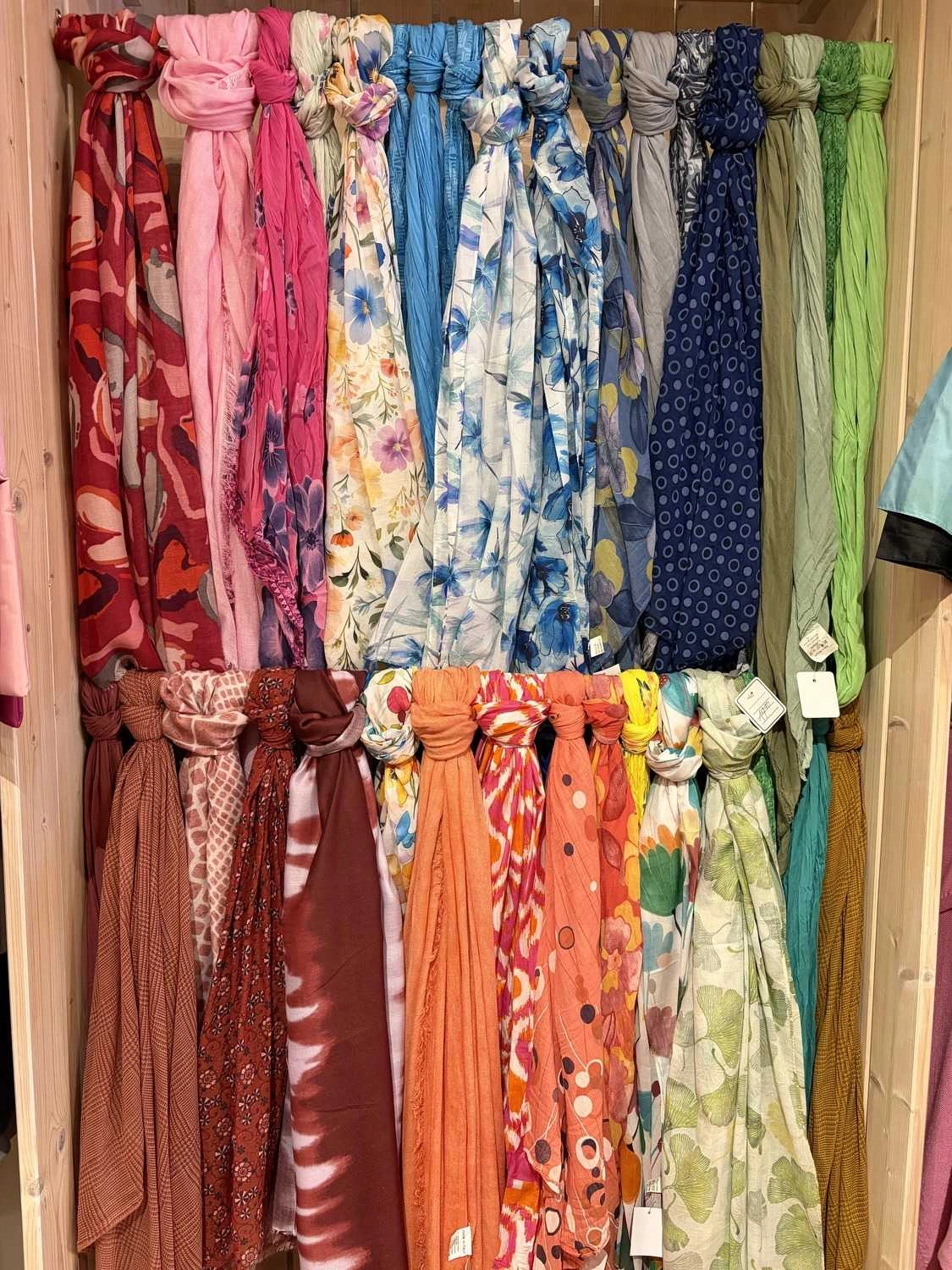 Mode & Handarbeitsdiele - Wolle, Kunsthandwerk, Mode und Accessoires - Bunte Schals mit verschiedenen Mustern und einfarbigen Varianten hängen senkrecht an einem Kleiderständer.