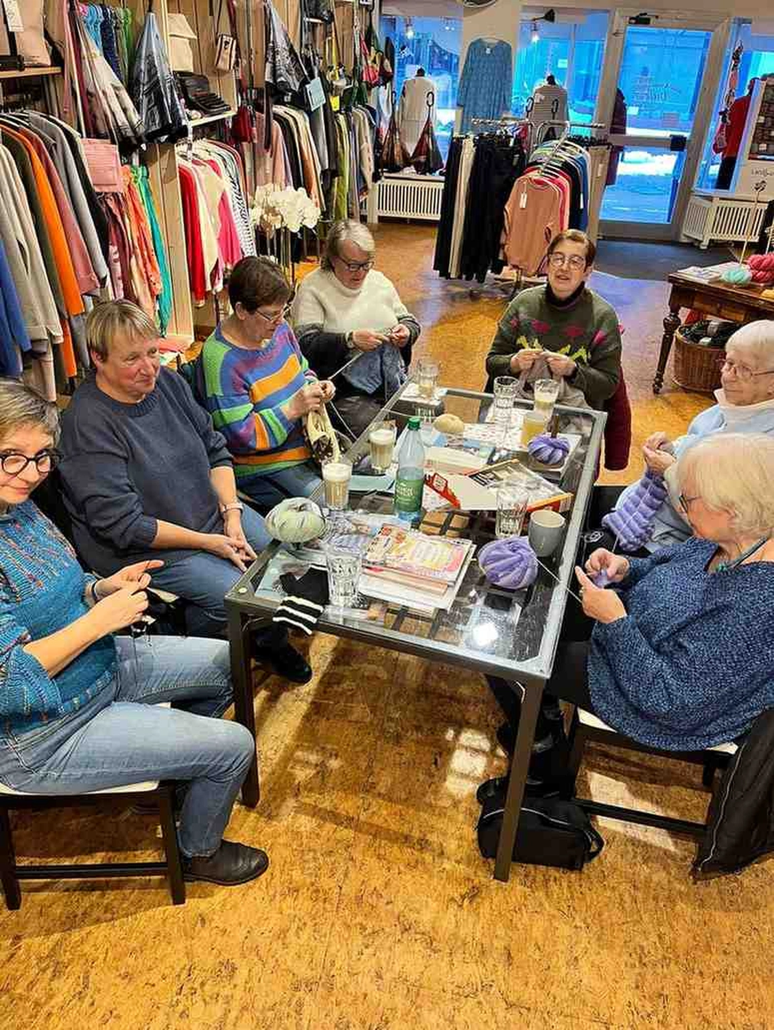 Mode & Handarbeitsdiele - Wolle, Kunsthandwerk, Mode und Accessoires - In einem hell erleuchteten Bekleidungs- und Wollgeschäft sitzen einige Frauen um einen Glastisch und sind in ihre Strickprojekte vertieft. Verschiedene Kleidungsstücke und Kleiderständer mit farbenfroher Kleidung füllen den Hintergrund des gemütlichen Raumes.